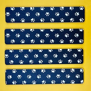 May include: Four rectangular nail files with a navy blue background and a repeating pattern of white paw prints. The files are arranged horizontally against a yellow background.