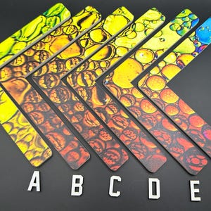 May include: Six L-shaped wooden rulers with abstract designs in yellow, orange, and red hues. Each ruler has a unique pattern resembling bubbles or cells. White letters A through F are at the bottom.