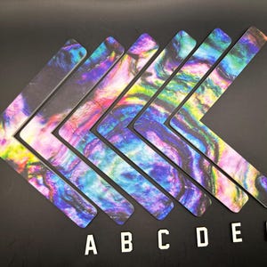May include: Seven L-shaped decorative pieces with a colorful, swirling pattern in shades of purple, blue, green, and yellow. The pieces are arranged in a row on a black surface, with the letters A through F below them.