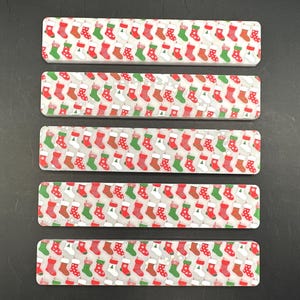May include: Five rectangular Christmas-themed decorations. Each features a repeating pattern of red, green, and white Christmas stockings on a light gray background. The decorations have rounded corners.