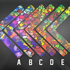 May include: A collection of six wooden boomerang-shaped items, each featuring a stained-glass design in vibrant colors. The boomerangs are arranged on a black surface, with letters A through F labeling each one.