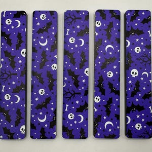 May include: Five rectangular bookmarks with a purple background and a repeating pattern of white skulls, crescent moons, stars, bats, and bones. The bookmarks have a Halloween theme.