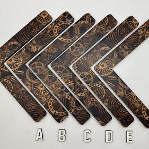 May include: Six brown and black steampunk-themed arrow-shaped decorative pieces. Each piece features a repeating pattern of gears and cogs. The letters A, B, C, D, E, and F are placed below the arrows.