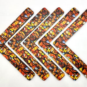May include: Six decorative, L-shaped items with a stained-glass pattern in shades of orange, yellow, and blue. The items are arranged in a chevron pattern against a white background. The design resembles a mosaic.