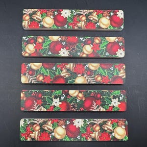 May include: Five rectangular Christmas-themed decorative items. Each features a repeating pattern of red and gold ornaments, bells, poinsettias, and holly leaves on a green background. The items are arranged on a dark surface.