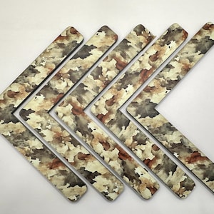 May include: A set of six L-shaped wooden shelf brackets. The brackets feature a camouflage pattern in shades of brown, beige, and gray. The brackets are arranged in a diagonal pattern against a white background.