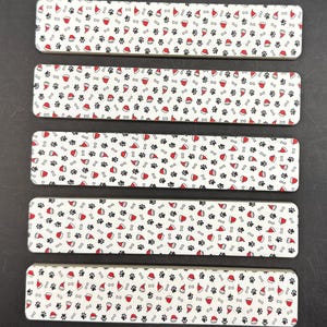 May include: Five rectangular, white decorative items with a repeating pattern of black paw prints, red Santa hats, and white bones. The items are arranged in a row, showcasing a festive, pet-themed design.