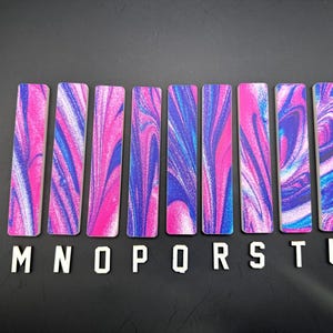 May include: A collection of eight rectangular bookmarks with a vibrant pink, blue, and white swirled pattern. The letters "M", "N", "O", "P", "R", "S", "T", and "U" are arranged below the bookmarks on a black surface.