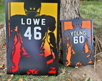 May include: Two black metal silhouette signs of football players with their names and numbers. The sign on the left has a yellow, orange, and red background and says "LOWE 46". The sign on the right has an orange background and says "YOUNG 60".