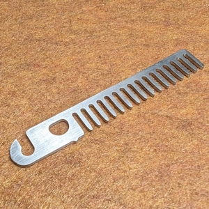 May include: A silver-colored metal tool with a comb-like edge and a hook on one end. The tool has a rectangular shape with a series of evenly spaced teeth along one side. A circular hole is near the hook. The tool is on a brown surface.