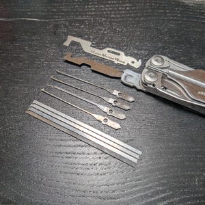 May include: A multi-tool with a brown handle and silver tools, including a saw, knife, and screwdriver. Several silver lock-picking tools and a metal ruler are also visible. The text "Machine Works" is on one of the tools.