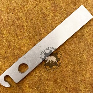 May include: A silver metal tool, rectangular with a curved hook and a hole. The tool has the text "MISHKA MACHINE WORKS" and a bear logo. It rests on a textured, brown surface. A useful accessory.