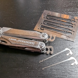May include: A silver Leatherman Wave+ multi-tool with the brand name visible. Beside it are two silver lock picks and a metal template with cut-out lock pick shapes. The items are on a dark surface.