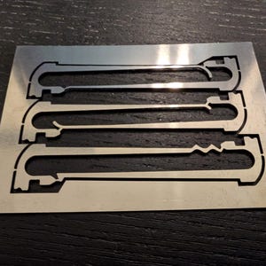 May include: A rectangular, brushed metal plate featuring three cut-out designs. Each design has a U-shaped end connected by a straight or zig-zag line. The plate is set against a dark, textured surface.