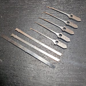 May include: A set of seven silver-coloured metal lock picking tools arranged in a diagonal pattern. The collection includes five slender picks with distinct hook and rake tips featuring circular holes in the handles, and two flat tension wrenches.