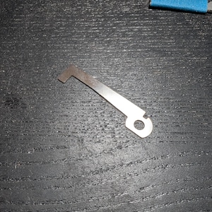 May include: A flat, silver-coloured metal component featuring an elongated, tapered body with a small circular hole at one end and a perpendicular, hook-like tab at the opposite end. The piece has a smooth, metallic finish and a distinct geometric shape.