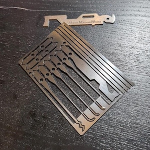 Pode incluir: Um conjunto de ferramentas de metal de precisão, incluindo uma folha retangular com várias formas recortadas e uma ferramenta separada em forma de chave. O texto "Mishka Machine Works" é visível na ferramenta em forma de chave. As ferramentas são feitas de metal prateado.