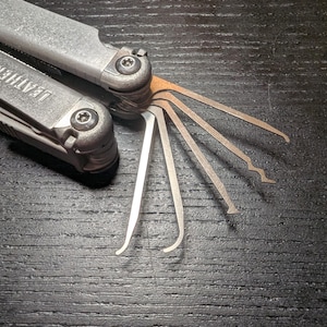 May include: A silver-coloured Leatherman multi-tool with several thin, metallic lock-picking implements extended from the handle. The tool features a brushed metal finish with the brand name Leatherman embossed on the side of the handle.