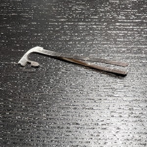 Op de afbeelding: Een klein, zilverkleurig metalen gereedschap met een gebogen haak aan één uiteinde en twee platte, parallelle pinnen die zich van de andere kant uitstrekken. Het gereedschap ligt op een donker, getextureerd oppervlak.