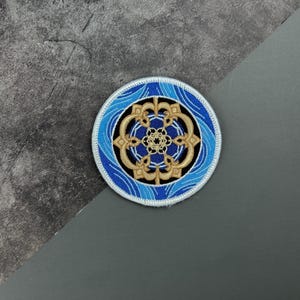 May include: A blue and gold embroidered patch with a circular design. The center of the patch features a gold mandala with intricate details. The mandala is surrounded by a blue wave pattern.
