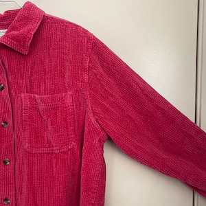 Puede incluir: Una camisa de pana rosa brillante con cuello, bolsillo en el pecho y botones. La camisa de manga larga está hecha de tela acanalada y está colgada en una percha.