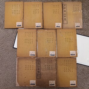 May include: A collection of ten antique books with brown covers and Japanese text. The books are stacked in two rows, with the top row containing five books and the bottom row containing five books. Each book has a small white label with a number on it.