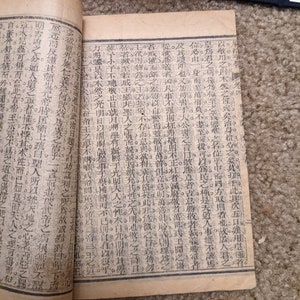 May include: An open book with pages filled with black text printed on a cream-colored background. The book is bound in brown paper and has a worn, aged appearance.