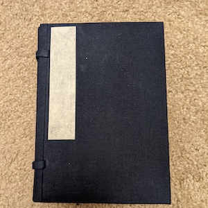 May include: A black hardcover journal with a beige tape bookmark. The journal has a textured cover and a simple design.