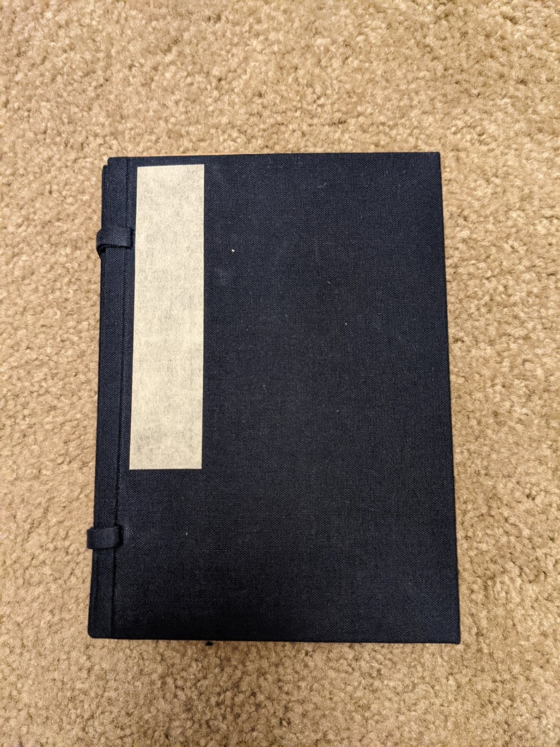 May include: A black hardcover journal with a beige tape bookmark. The journal has a textured cover and a simple design.