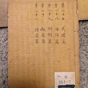 May include: A tan-colored book with black text on the cover. The text reads "第二十七 武備篇", "芽二十八海防篇", "券二十九", "麥三十", "刑制篇", "赦看篇", "岸三十一弭盗篇". The book has a black label on the bottom right corner with the text "No. H 354-7".