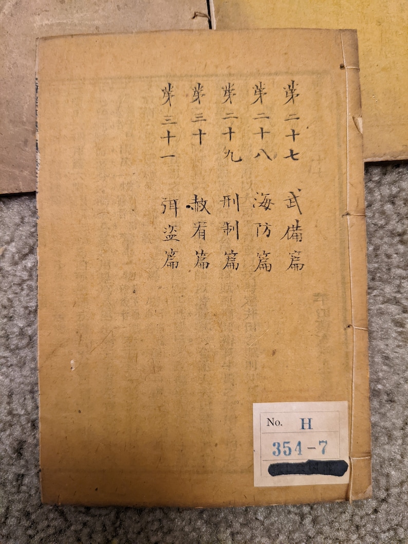 May include: A tan-colored book with black text on the cover. The text reads "第二十七 武備篇", "芽二十八海防篇", "券二十九", "麥三十", "刑制篇", "赦看篇", "岸三十一弭盗篇". The book has a black label on the bottom right corner with the text "No. H 354-7".