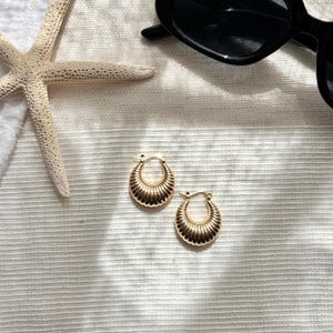Peut inclure: Une paire de boucles d'oreilles dorées à créoles avec un motif côtelé, à côté de lunettes de soleil noires et d'une étoile de mer sur une surface blanche texturée. Les boucles d'oreilles ont un style classique et élégant.