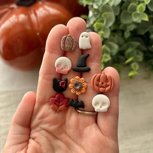 May include: A hand holds various Halloween-themed charms. The charms feature pumpkins, ghosts, skulls, a witch's hat, a black cat, a sunflower, a leaf, and a bat. A large orange pumpkin is visible in the background.