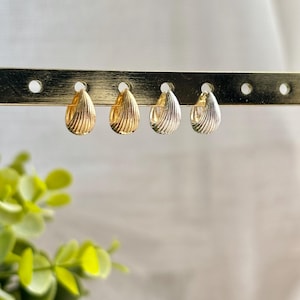 May include: Four pairs of teardrop-shaped earrings with a ridged design are displayed on a gold-coloured earring stand. Two pairs are gold-toned, and two are silver-toned. The earrings are hanging in a row against a white background.