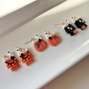 Peut inclure: Trois paires de boucles d'oreilles à tige représentant des chiens blancs. Une paire montre des chiens avec des couvertures rayées orange et des guirlandes, une autre avec des citrouilles orange, et la troisième avec des tenues noires et des étoiles jaunes, toutes avec des tiges dorées.