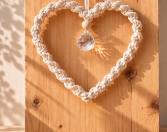Puede incluir: Un adorno de pared de macramé en forma de corazón hecho de cuerda de algodón trenzado color blanco roto. Una cuenta de madera se sitúa en la parte superior central, de la que cuelga un cristal transparente, facetado y en forma de gota.