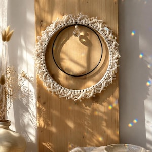 Könnte beinhalten: Ein kreisförmiger Wandbehang aus cremefarbener Makramee-Kordel, die um einen großen äußeren Ring und einen kleineren schwarzen inneren Ring geknotet ist. Eine klare, facettierte Kristallperle hängt mittig am oberen Rand des inneren Rings.