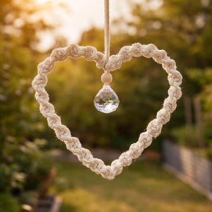 Könnte beinhalten: Ein herzförmiges Makramee-Ornament aus cremefarbener gedrehter Kordel, das an einer passenden Schnur hängt. Eine kleine Holzperle sitzt über einem klaren, facettierten Kristallanhänger, der mittig im herzförmigen Rahmen hängt.