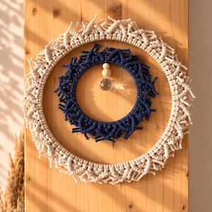 Könnte beinhalten: Ein kreisförmiger Makramee-Wandschmuck bestehend aus einem großen äußeren Ring aus cremefarbener Kordel und einem kleineren inneren Ring aus dunkelblauer Kordel. Das Design zeigt komplexe Knotenmuster und ein zentrales Element mit zwei Holzperlen.