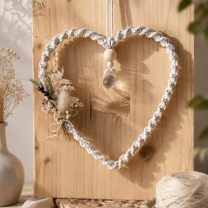 Può includere: Una decorazione da parete in macramè a forma di cuore realizzata in corda color crema, con una perla di legno centrale e un ciondolo in cristallo trasparente sfaccettato. Un piccolo bouquet di fiori secchi color crema, beige e viola è fissato sul lato sinistro.