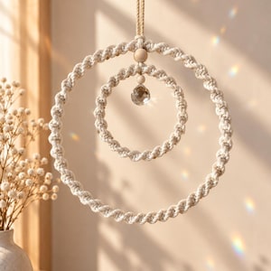 May include: A hanging macrame ornament featuring two concentric circles made of off-white twisted cotton cord. A single wooden bead and a clear, multifaceted crystal prism are suspended from the top centre of the inner circle by a cord.