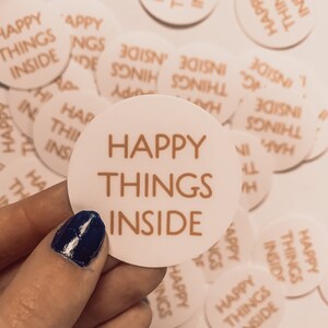 May include: A collection of white round stickers with the text "Happy Things Inside" printed in gold. The stickers are scattered on a white surface.