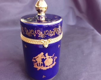 May include: A blue and gold porcelain trinket box with a lid. The box is decorated with a gold floral design and a scene of two figures in a garden. The box is approximately 4 inches tall.
