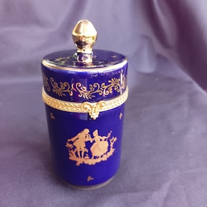 May include: A blue and gold porcelain trinket box with a lid. The box is decorated with a gold floral design and a scene of two figures in a garden. The box is approximately 4 inches tall.