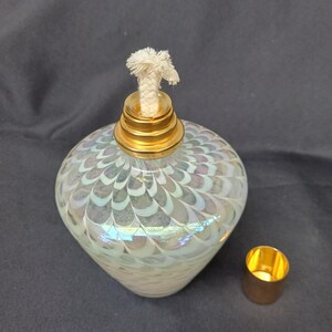 May include: A pear-shaped, iridescent glass oil lamp with a gold-colored metal top and wick. The lamp has a decorative pattern and a separate gold-colored metal cap. The lamp is designed for use with oil.