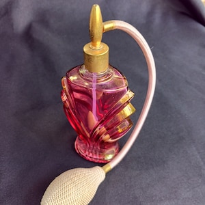 May include: A vintage glass perfume bottle with a pink and gold design and a long, thin, pink spray nozzle. The bottle has a decorative, geometric design.