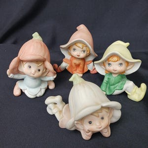 May include: Four ceramic figurines of flower fairies in various poses. Each fairy has a flower-shaped hat and is dressed in pastel colors, including pink, orange, green, and white. The figurines have detailed facial features.