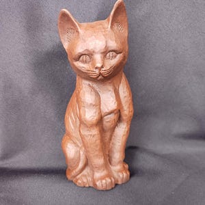 May include: A brown, carved wooden cat figurine in a seated position. The cat has detailed facial features, including eyes, a nose, and whiskers. The figurine is a solid color and has a textured surface.