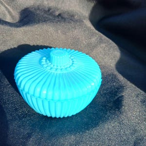 May include: A bright turquoise, lidded trinket box. The box is round with a ribbed, fluted design on the sides and a decorative top. The box is made of a solid, opaque material and sits on a dark surface.