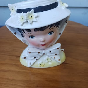 May include: A vintage ceramic planter shaped like a girl's head, wearing a white hat with a black band and floral accents. The planter has a yellow base with a white bow and polka dots. The girl has blue eyes and red lips.